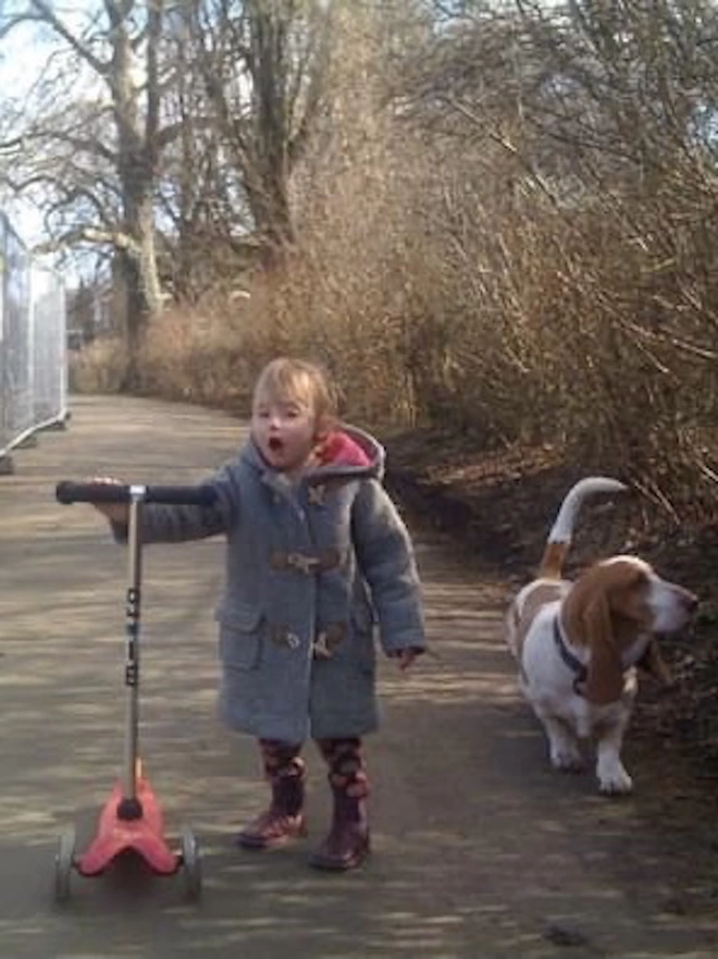 Piper as a 5 year old in a grey duffel coat pushing her pink scooter next to her basset hound dog bunty