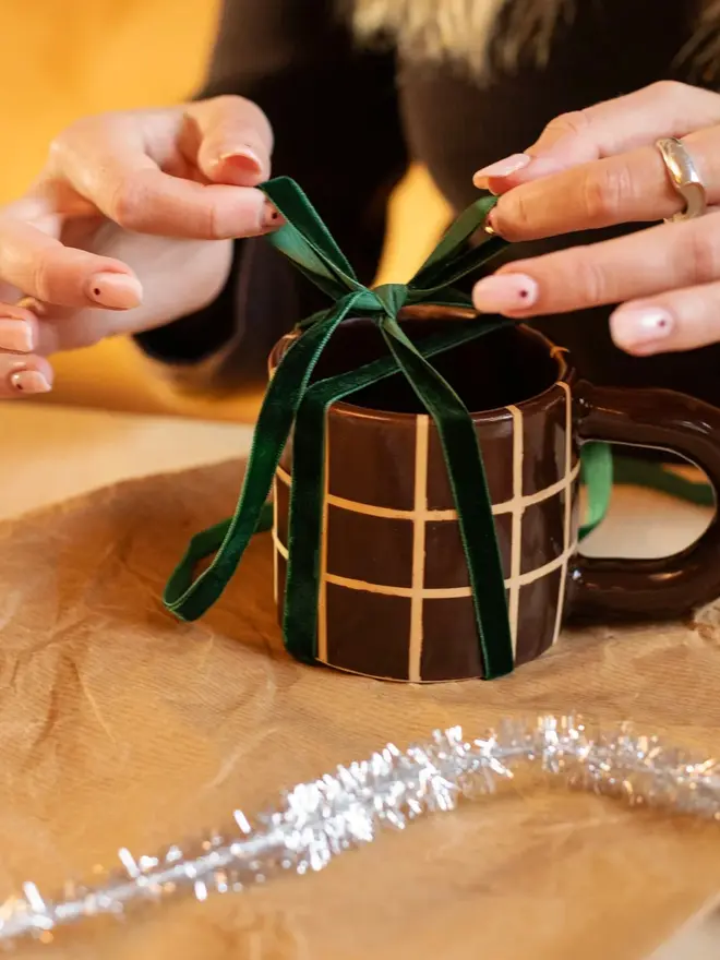 Grid Chunky Mug, a colourful mug with a grid pattern on a white table top surface. It is being held above some wrapping paper. 