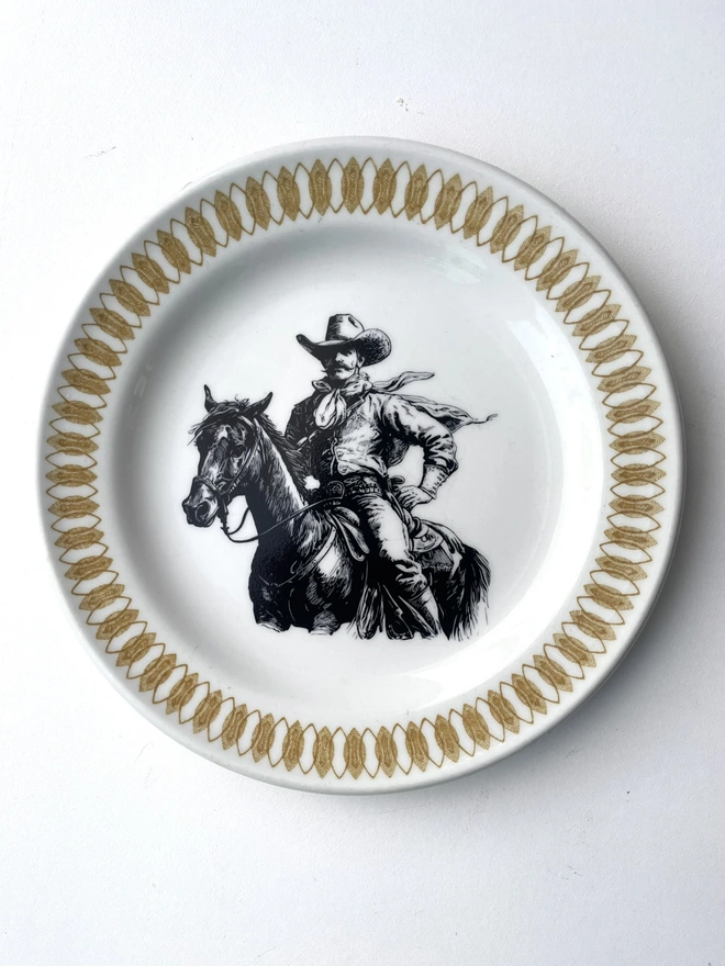 vintage plate hanging on a white wall, it has a decorative border, with a printed vintage illustration of a cowboy in the middle 
