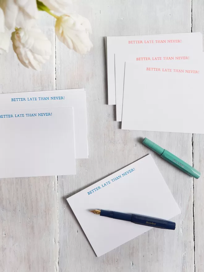 Sets of the Better Late Than Never Cards in orange and blue on a wooden table with two fountain pens.