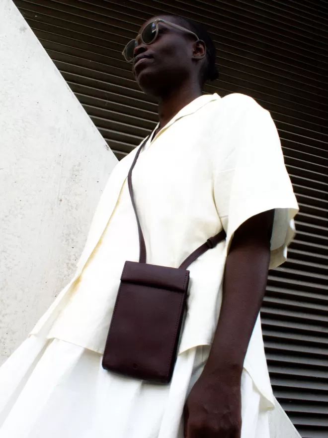 Oxblood Phone Bag worn beautifully worn on a model of darker complexion seen from the bottom up. She is wearing a completely neutral cream oversized shirt and skirt against an industrial wall and metal shutter.