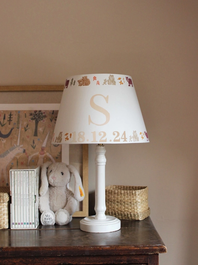 Cozy corner of a girls bedroom, featuring a wooden dresser and white table lamp paired with a personalised kids lampshade adorned with a bears pattern and the letter 'S'. A framed artwork hangs behind the lampshade featuring a unicorn and mythical setting. 