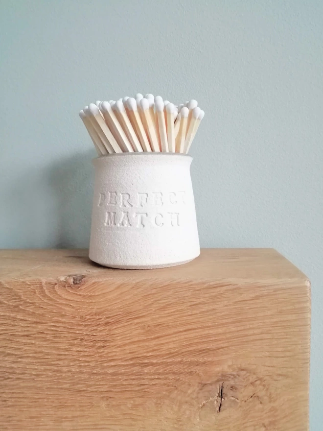 Match pot sat on wooden fireplace. Pot caontains white tip matches with 'Perfect Match' stamped on the outside 