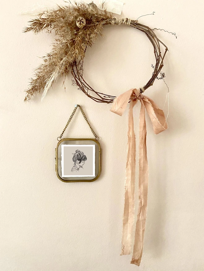 a tiny portrait hand drawn hung in a glass frame with a brass border hung next to a hand made wreath on a peach wall