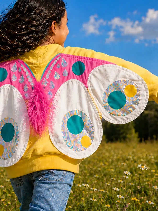 A little girl, approx 6 yrs old is running through a field wearing a yellow sweatshirt with butterfly wings attached to the back