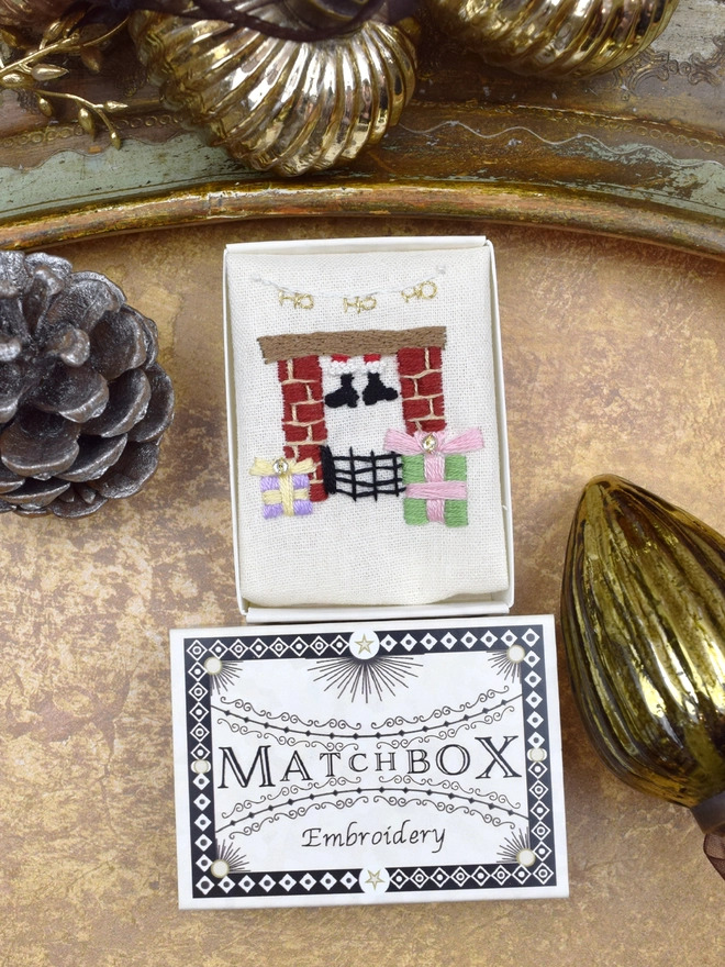 The fireplace embroidery, inside a Matchbox displayed on a gold background with glass baubles.