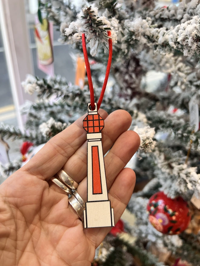 Small white and orange wooden tree decoration in the shape of Margate Lido sign. It is hanging by a red thread on a white fake Christmas Tree with other decorations. There is a hand cradling the decoration in the shot.