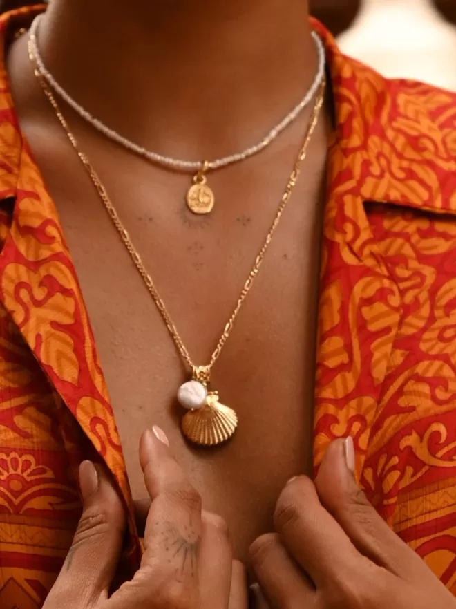 Model wearing gold vermeil long shell pendant with baroque pearl layered with pearl bead necklace and small oval charm