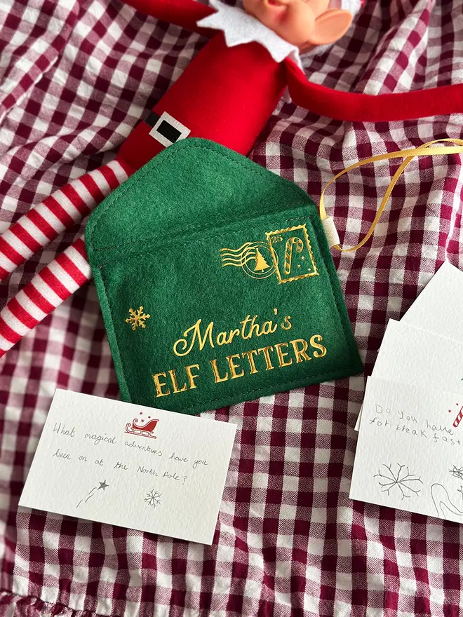 A handmade green Christmas envelope with a festive card tucked inside, written with a note to the elves, lays on a gingham cloth next to a Christmas elf toy.