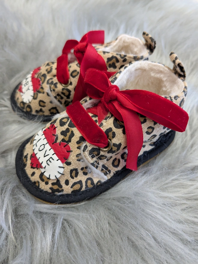 A pair of leopard print baby booties, lined with fur and with red velvet ribbon laces. A red heart with a white scroll adorns each booty. One says BABY, the other, LOVE in black stitches