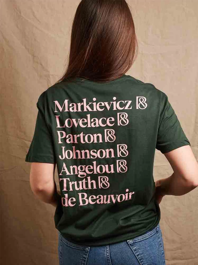 Model is showing the back of the forest green feminist icon t-shirt. The names of our chosen icons, Markievicz, Lovelace, Parton, Johnson, Angelou, Truth, and De Beauvoir printed in pale pink 