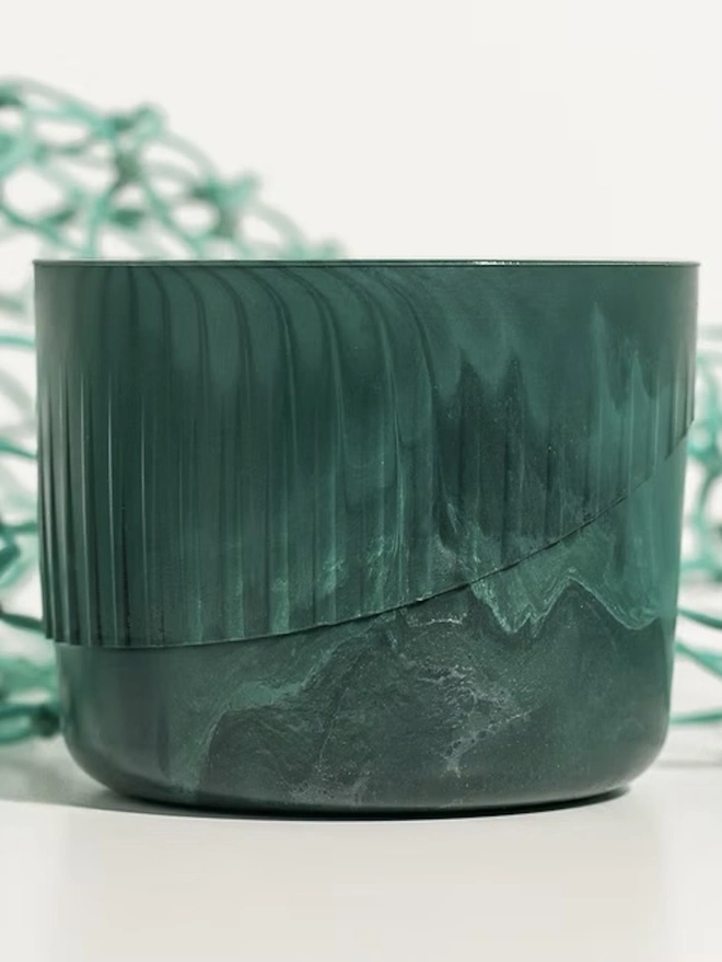 Ocean Plastic Plant Pot Small, a green recycled plant pot sitting on a white surface against a plain backdrop. It is accompanied by some recycled fishing nets.  