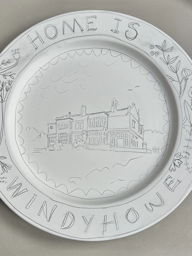 A sketch of a house on a hill in pencil prior to painting and firing. "Home is Windyhowe" written around the rim, with the large house inset. a botanical border between the words