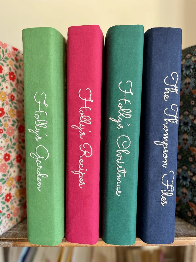 Close-up of 4 fabric covered ring binders on a bookshelf with green, pink, dark green and navy blue spines showing, all embroidered with personalisation in thick white thread, one saying 'The Thompson Files', another 'Holly's Garden'.