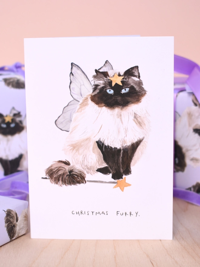 Liz Temperley Christmas Furry Card. On the front is a fluffy cat wearing wings with a star wand and star on its head. Behind is the matching wallpaper.