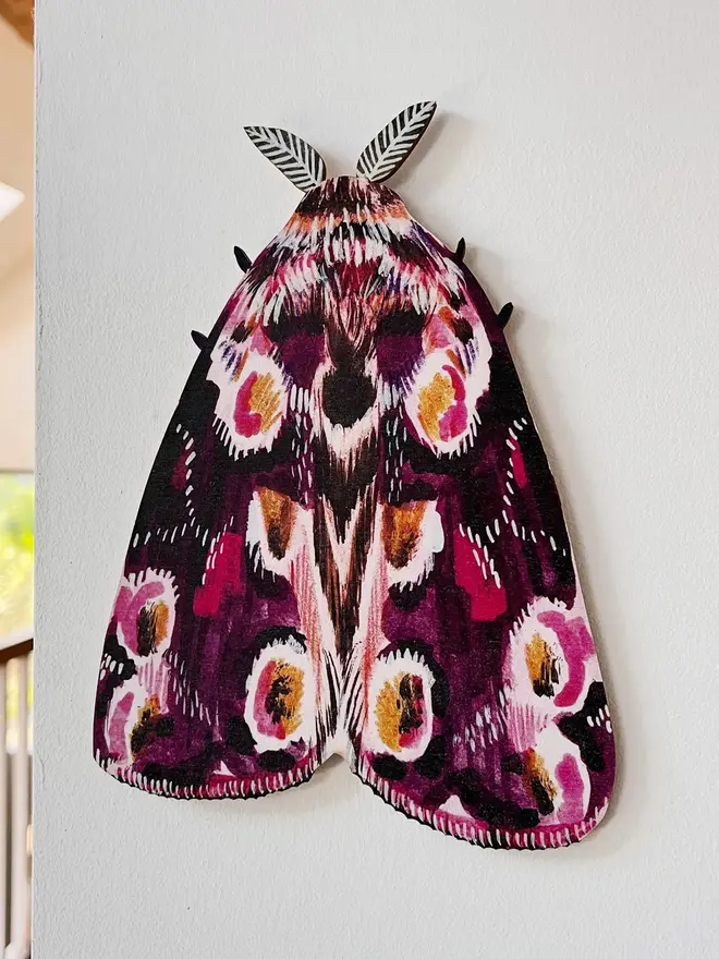 large wooden moth wall decoration. large pink wooden moth decoration hung on plain white wall.