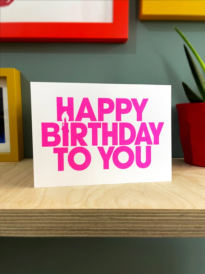 Bright pink text screen printed on a white landscape card, the bold capital font contains a candle created in the negative space of the words. The card sits on a plywood shelf with coloured frames showing at the edges and part of a plant in shot. 
