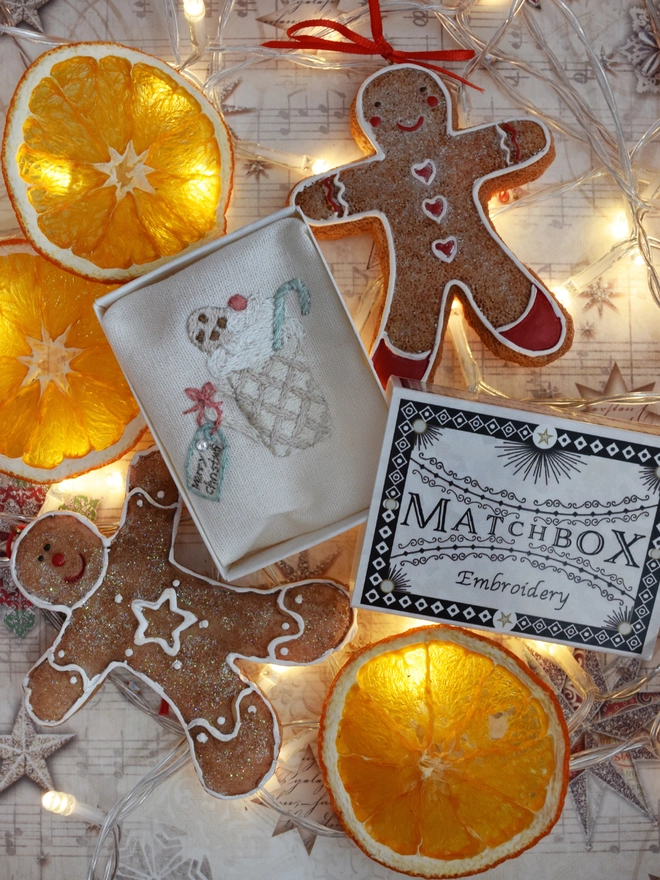 The Mug of Cocoa embroidery displayed in its matchbox.  Surrounded by dried orange slices, gingerbread decorations and fairy lights. 