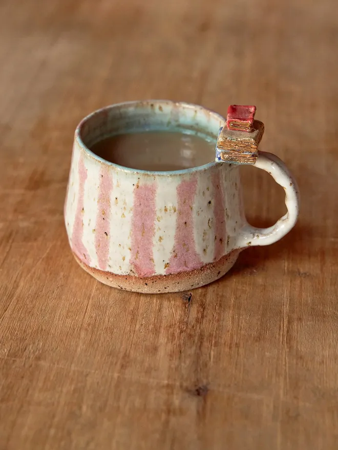 Pink and white striped ceramic mug with book detail on the handle, pictured on a wooden table