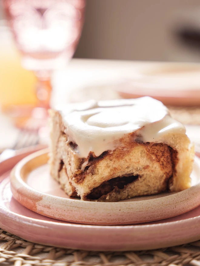 Close up of a cinnamon bun on a pink plate, covered in vanilla frosting