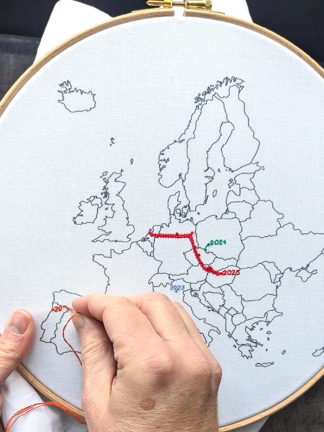Fabric with Europe printed on it, in a wooden embroidery hoop, with hands stitching Portugal.