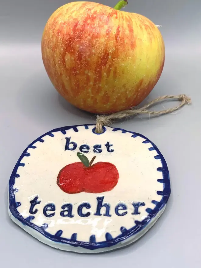 'Best Teacher' Handcrafted Ceramic Ornament, a ceramic apple ornament sitting on a plain white surface joined by an apple. 