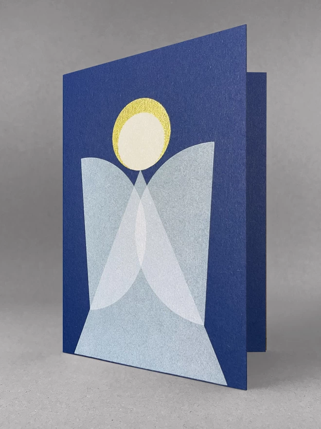 Slightly open, a geometric angel christmas card design, on blue card, white overlapping wings and a gold halo. Sits in a light grey studio