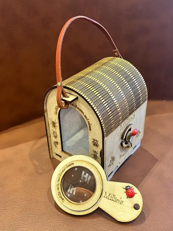 Wooden bug hotel for kids with engraved insect illustrations, mesh viewing window, and leather carry handle, displayed on a table with a matching wooden magnifying glass.