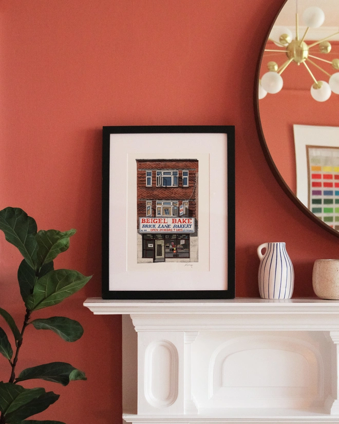 A framed print featuring an illustration of Beigel Bake displayed on a white mantelpiece against a terracotta-colored wall. 