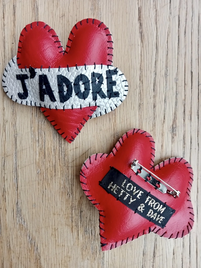 Two red heart brooches, one face up saying J'ADORE in black lettering across a white scroll. The other is face up, showing off the brooch back, and Love From Hetty & Dave label