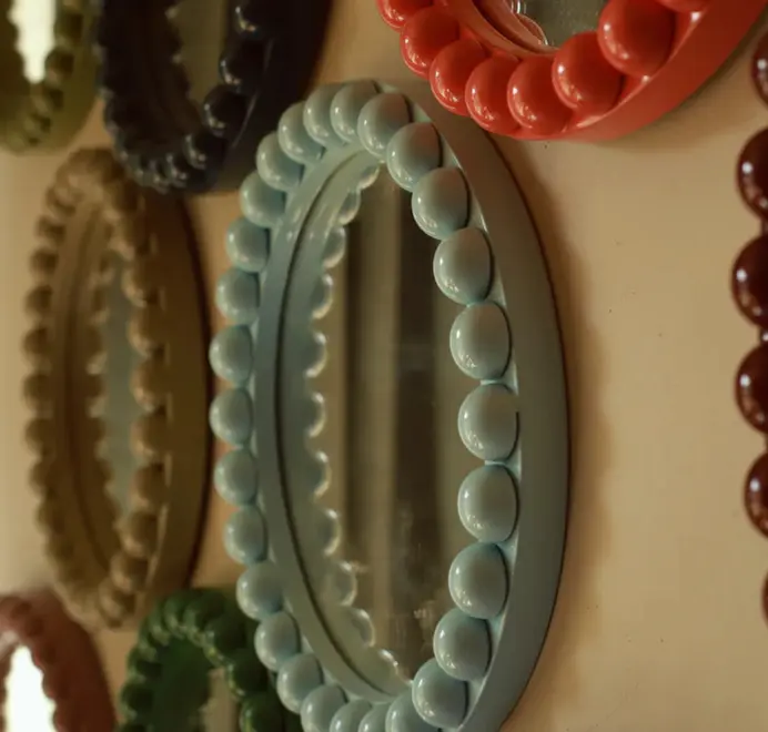 Many round oversized bobbin mirrors, in different colours and sizes with a decorative bobbin-style wooden frame, hanging on a white wall.