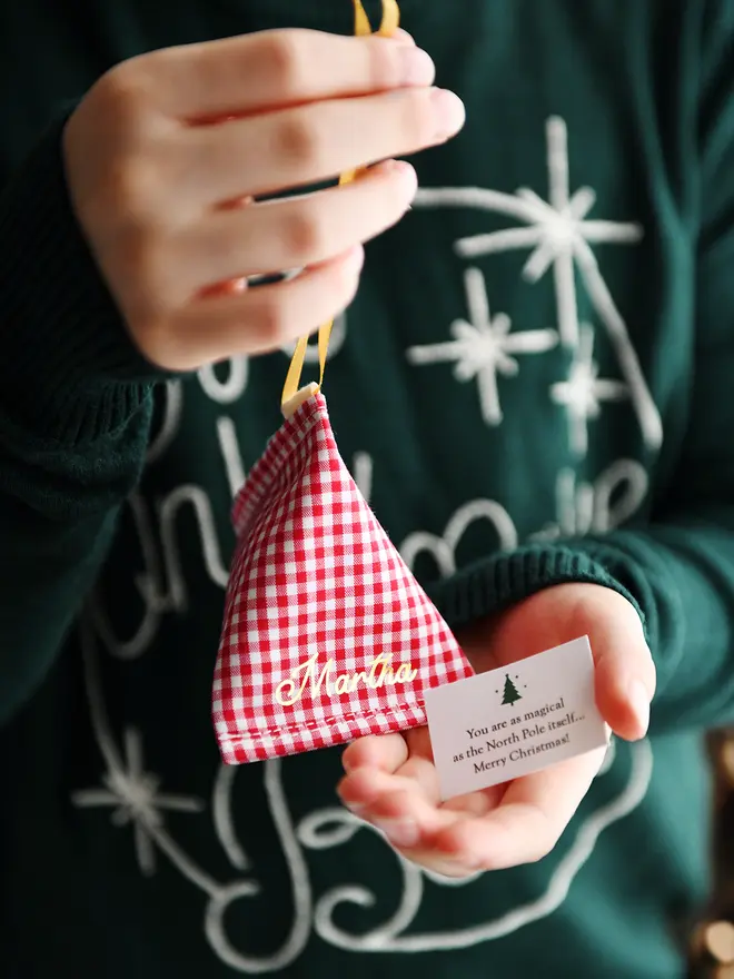 A child wearing a green Christmas jumper holds a red gingham personalised fabric fortune cookie with the name “Martha” in gold, revealing a tiny wish card inside that reads “You are as magical as the North Pole itself… Merry Christmas!”