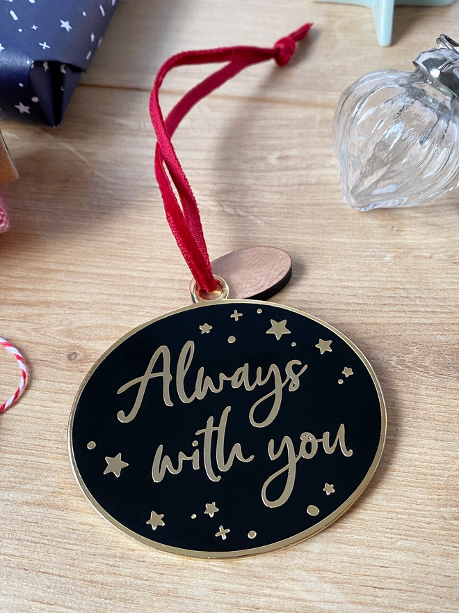 A black and gold enamel Christmas decoration, with the words “Always With You” surrounded by gold stars, lays on a wooden desk.