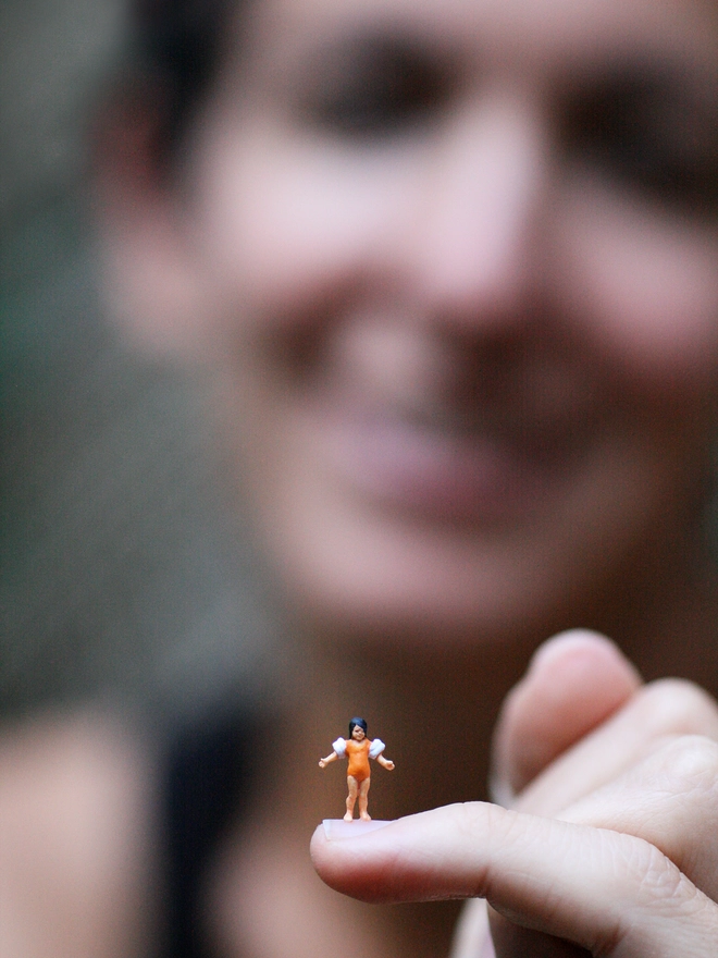 A miniature figurine of a little girl in armbands, perched on the artist’s baby finger 