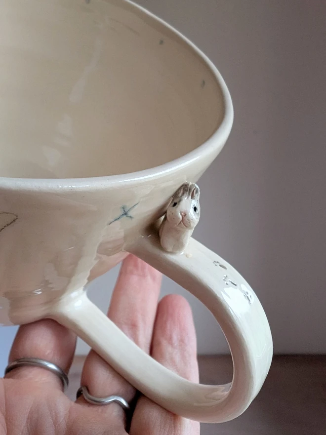 Close up of a tiny grey and white bunny rabbit on the handle of a white cup with tiny pawprints 