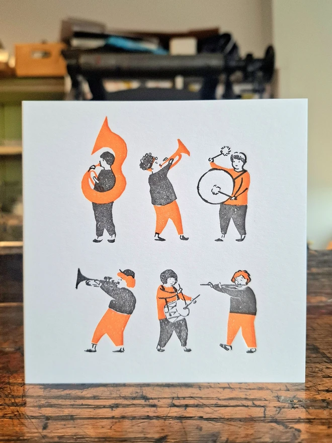 A greetings card standing open on a wooden surface, with an orange and black illustration of small figures playing instruments on the front.
