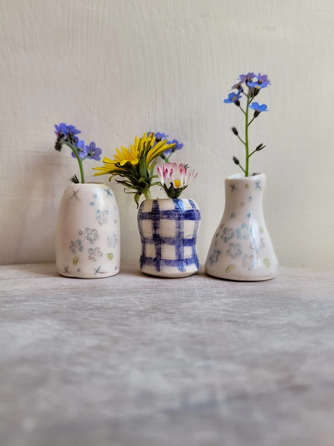 3 miniature ceramic vases 2 with hand painted blue flowers and 1 with a blue check pattern in a row on a wooden surface with forget me not dandelion and daisy flowers inside