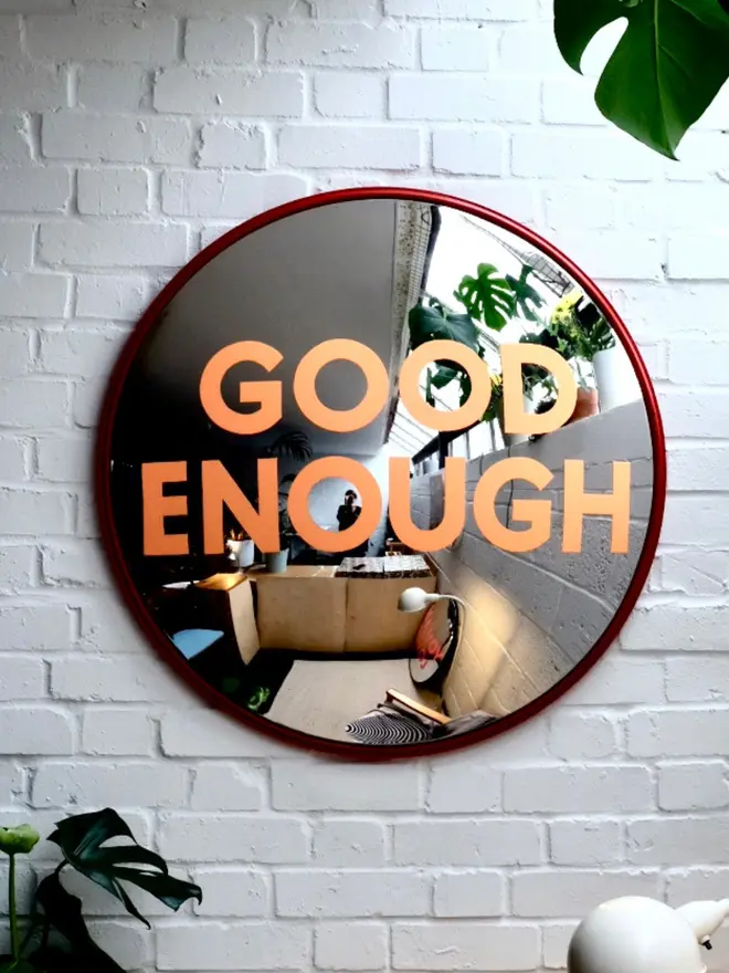 Round convex mirror with 'Good Enough' hand painted in orange in a red round frame, on a white brick wall with foliage around