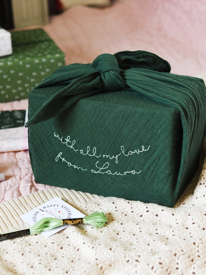 A gift wrapped in a deep green cotton fabric wrap that has been embroidered with a special message. 