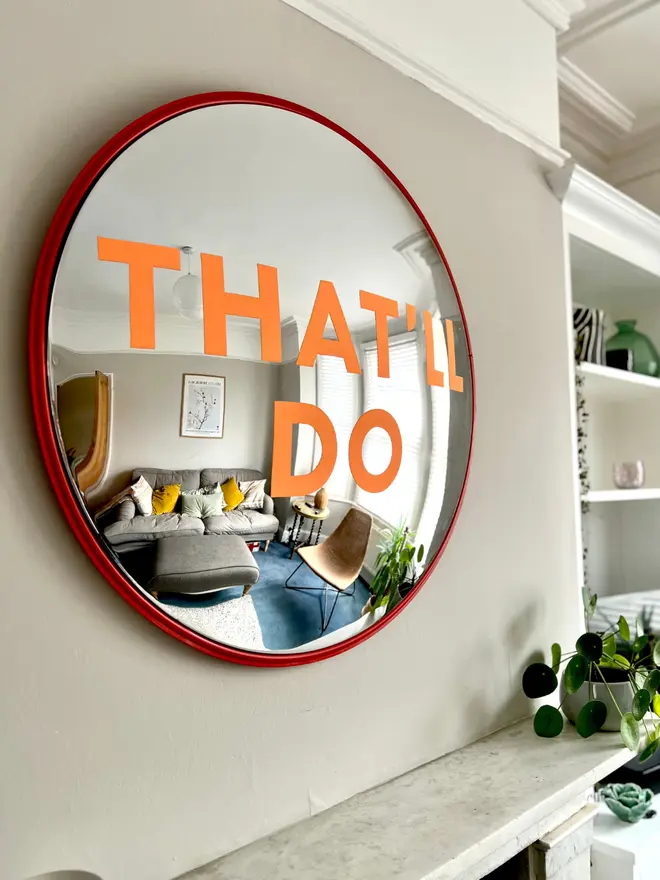 Round convex mirror with 'That'll Do' hand painted in orange in a red round frame, above a cream fireplace surrounded by house plants