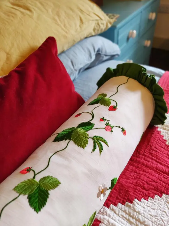 Close-up of strawberry plant embroidery on a cream bolster cushion with deep green gathered ruffle trim.