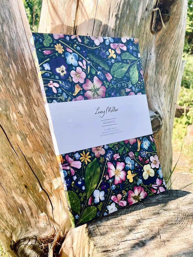 Eco-Friendly Notebook with Pressed Flower Design Cover, Staples Spine, 'Lucy Miller' Branded Belly Band, Leaning Against Tree Trunk