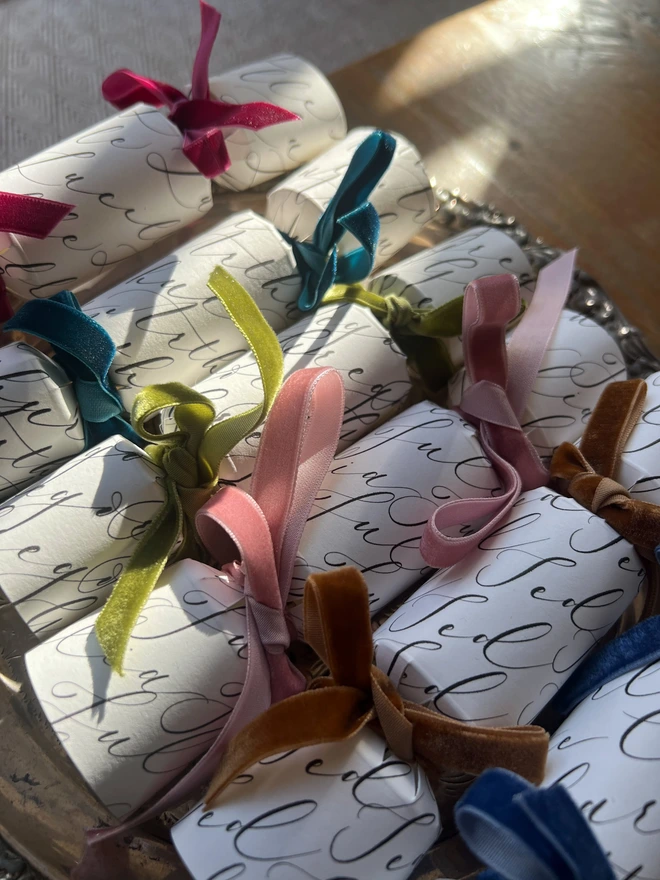 Selection of crackers personalised with calligraphy names tied with green, pink, mustard, blue, teal, rose, cerise velvet ribbon, laid on a silver tray on a wooden table 