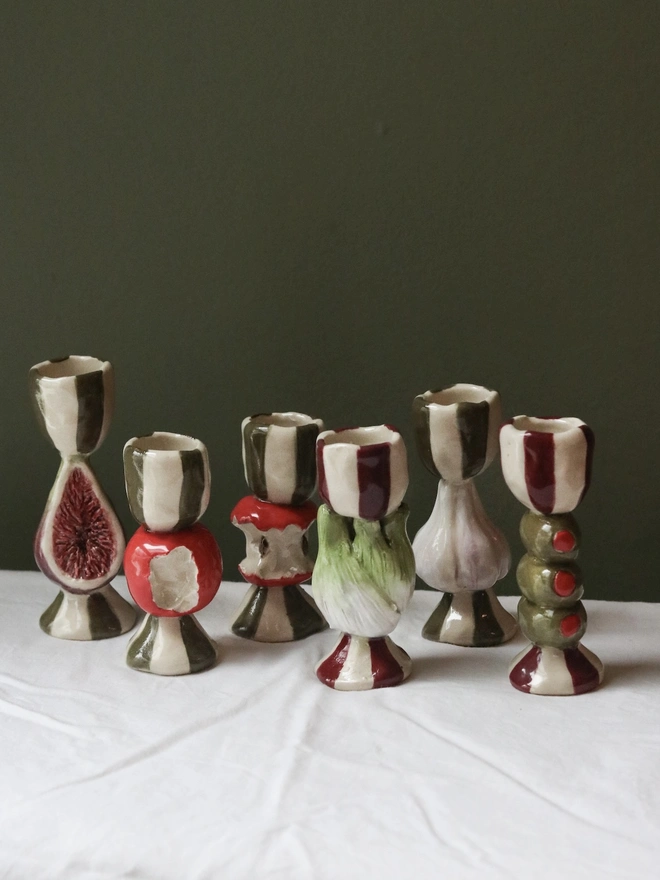 Candlesticks, a group of candle sticks on a white tablecloth. They have small emblems of fruits or vegetables on them. 
