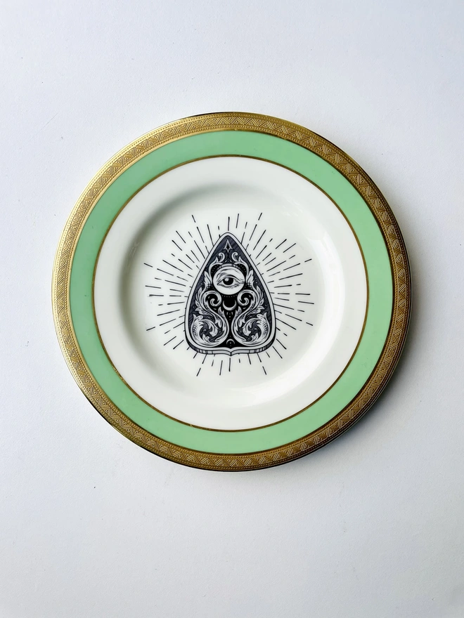 A vintage plate with a green and gold border, the middle of the plate has a vintage illustration of a ouija planchette