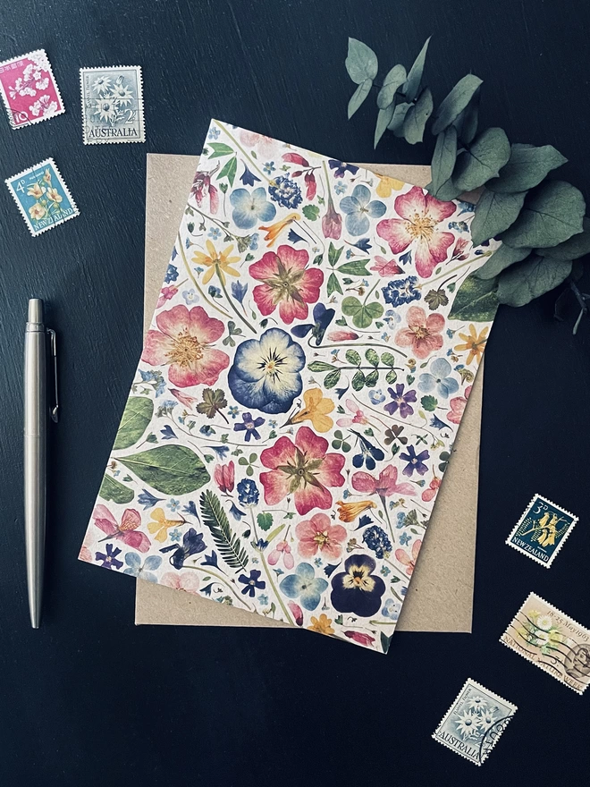 Floral Greetings Card with Colourful Pressed Flower Design - Brown Kraft Envelope - Dark Charcoal Coloured Desk - Colourful Floral Postage Stamps, Silver Ballpoint Pen, Dried Eucalyptus