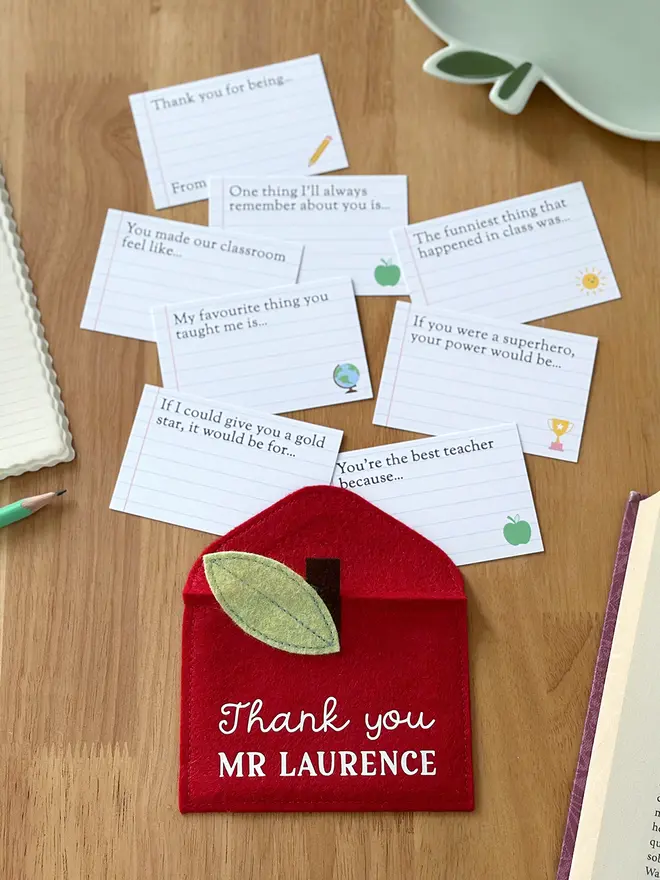 A handmade red felt apple-shaped envelope with "Thank you Mr Laurence" written on the front. The heirloom apple envelope surrounded by completed prompt cards with handwritten messages from a child, displayed on a wooden desk. It's a thoughtful personalised gift for teachers from children.