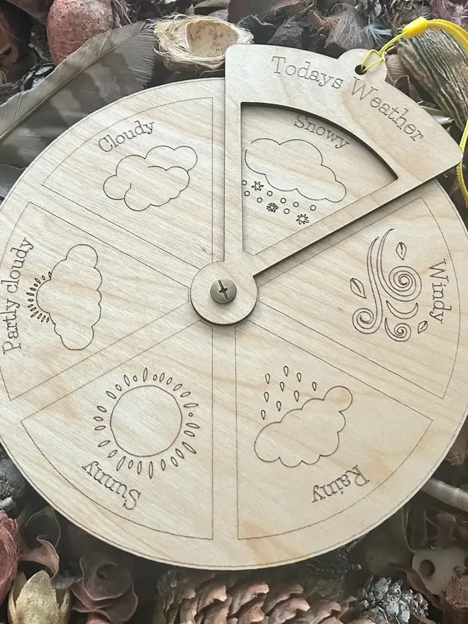 Extra large wooden weather wheel with engraved weather symbols for sunny, cloudy, rainy, windy, snowy, and partly cloudy, shown resting among pinecones and natural materials.