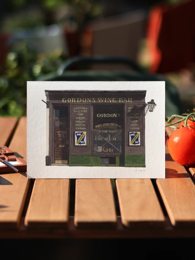 A watercolor print of Gordon's Wine Bar resting on a slatted wooden table outdoors next to a vine of fresh tomatoes.