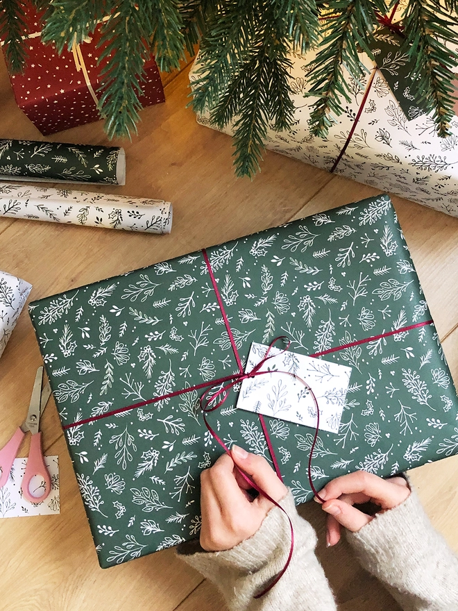 A gift wrapped in deep green wrapping paper with a Christmas botanical design lays on a wooden floor while a ribbon is being tied around it.